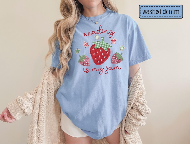 May include: A light blue washed denim t-shirt featuring a graphic of three strawberries with green gingham leaves and small stars. The text "reading is my jam" is printed in red cursive script above and below the central strawberry illustration.