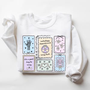May include: White sweatshirt featuring colorful illustrations related to radiology. The design includes images of a hand, a skeleton, and a radiology symbol, along with the text "radiology department" and "It's a good day to xray."