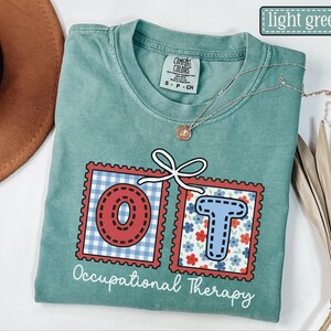 May include: Light green t-shirt with the text "Occupational Therapy". The letters "OT" are in red and blue postage stamp designs. A gold necklace with a pendant is on the shirt.