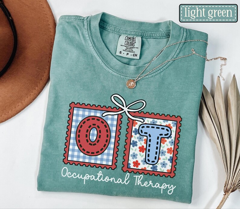 May include: Light green t-shirt with the text "Occupational Therapy". The letters "OT" are in red and blue postage stamp designs. A gold necklace with a pendant is on the shirt.