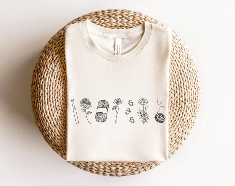 May include: A white t-shirt with a black line drawing of a crochet hook, yarn, and flowers.