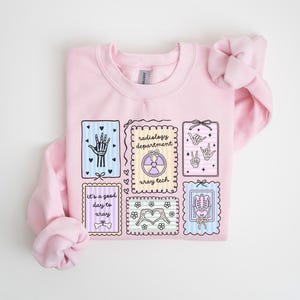 May include: A light pink sweatshirt featuring several illustrated rectangular designs. The designs include a hand, a radiology symbol, hand gestures, a skeleton, and the text "It's a good day to xray".