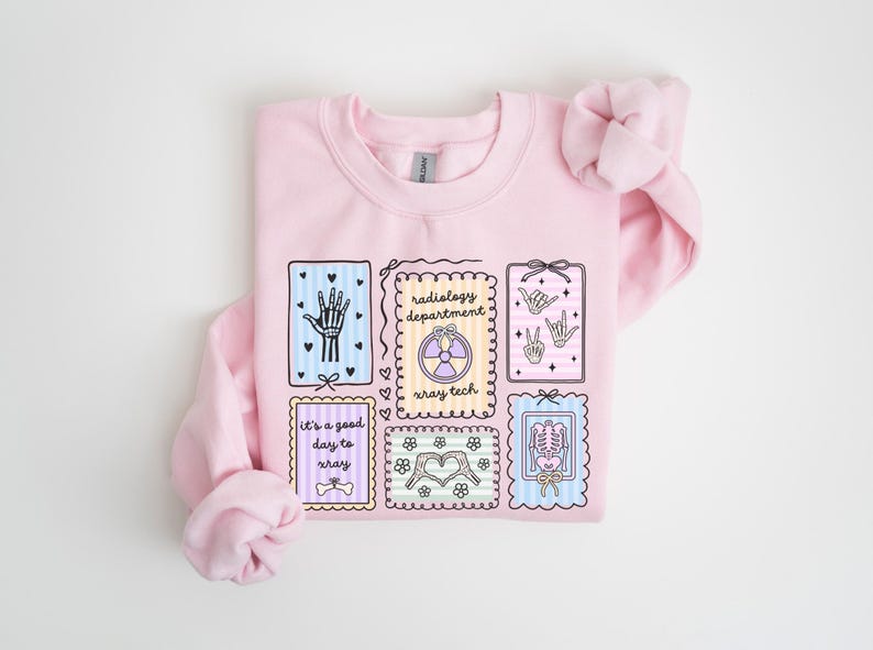 May include: A light pink sweatshirt featuring several illustrated rectangular designs. The designs include a hand, a radiology symbol, hand gestures, a skeleton, and the text "It's a good day to xray".