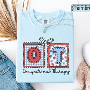 May include: Light blue t-shirt with the letters "OT" in red and blue, framed in a postage stamp design. The shirt also features the words "Occupational Therapy" in black script. A silver necklace and white shoes are also visible.