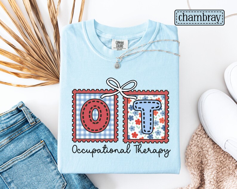 May include: Light blue t-shirt with the letters "OT" in red and blue, framed in a postage stamp design. The shirt also features the words "Occupational Therapy" in black script. A silver necklace and white shoes are also visible.