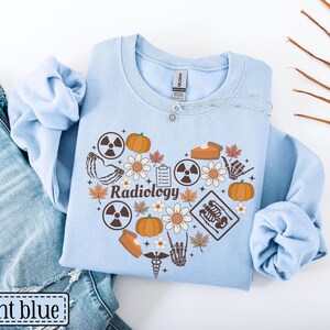 May include: A light blue crewneck sweatshirt featuring a heart-shaped graphic composed of pumpkins, flowers, autumn leaves, radiation symbols, a clipboard, a skeleton hand, a slice of pie, and a medical caduceus with the word Radiology in the center.