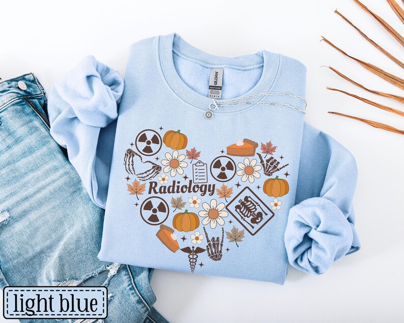 May include: A light blue crewneck sweatshirt featuring a heart-shaped graphic composed of pumpkins, flowers, autumn leaves, radiation symbols, a clipboard, a skeleton hand, a slice of pie, and a medical caduceus with the word Radiology in the center.
