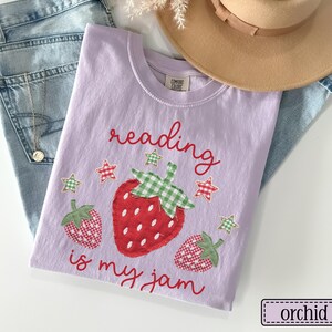 May include: A light purple t-shirt featuring the red text reading is my jam and a graphic of three strawberries with green and white chequered leaves. The strawberries have red and white chequered patterns. Small red and green stars surround the fruit.
