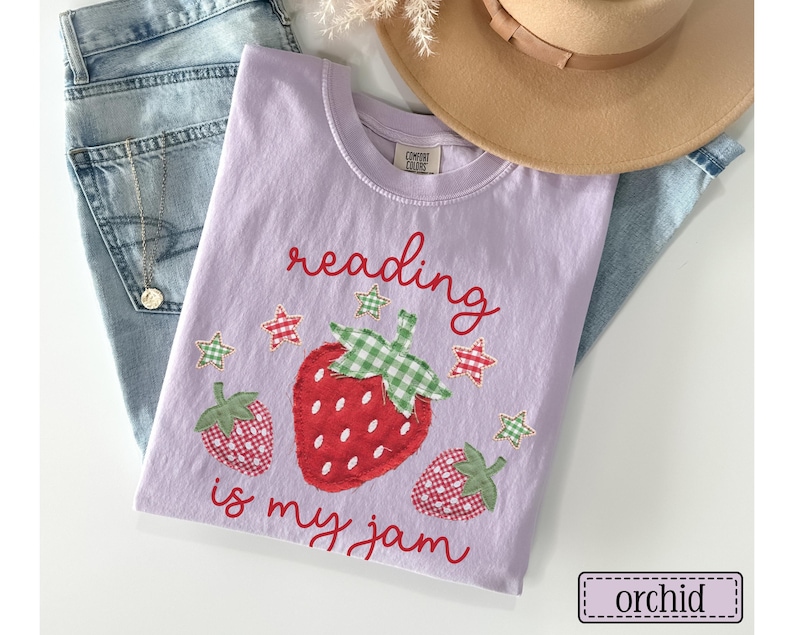 May include: A light purple t-shirt featuring the red text reading is my jam and a graphic of three strawberries with green and white chequered leaves. The strawberries have red and white chequered patterns. Small red and green stars surround the fruit.