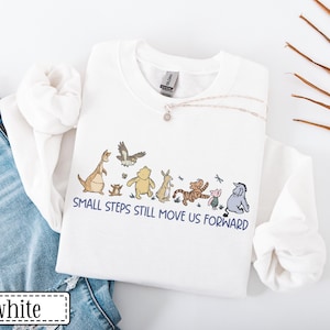 May include: White sweatshirt with a cartoon graphic of characters like a kangaroo, bear, tiger, and donkey. The text "SMALL STEPS STILL MOVE US FORWARD" is printed below. The sweatshirt is displayed with blue denim jeans.