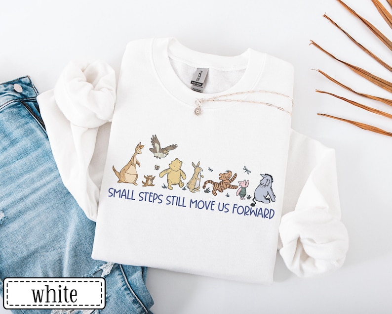 May include: White sweatshirt with a cartoon graphic of characters like a kangaroo, bear, tiger, and donkey. The text "SMALL STEPS STILL MOVE US FORWARD" is printed below. The sweatshirt is displayed with blue denim jeans.