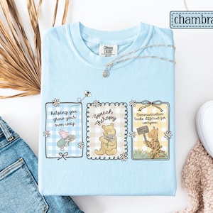 May include: Light blue Comfort Colors t-shirt featuring three framed illustrations with Winnie the Pooh characters. The frames contain the phrases: "helping you grow your own way," "Speech Therapy," and "Communication looks different for everyone."