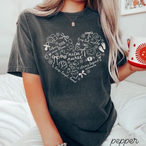 May include: Dark gray t-shirt with a white heart-shaped design featuring baby-related illustrations and the words "NICU nurse." The shirt also includes the phrases "helping little ones bloom," "tiny but mighty," and "tiny, tough, loved."