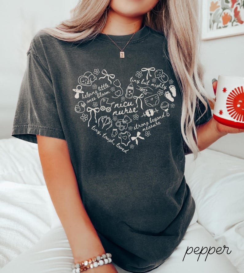 May include: Dark gray t-shirt with a white heart-shaped design featuring baby-related illustrations and the words "NICU nurse." The shirt also includes the phrases "helping little ones bloom," "tiny but mighty," and "tiny, tough, loved."