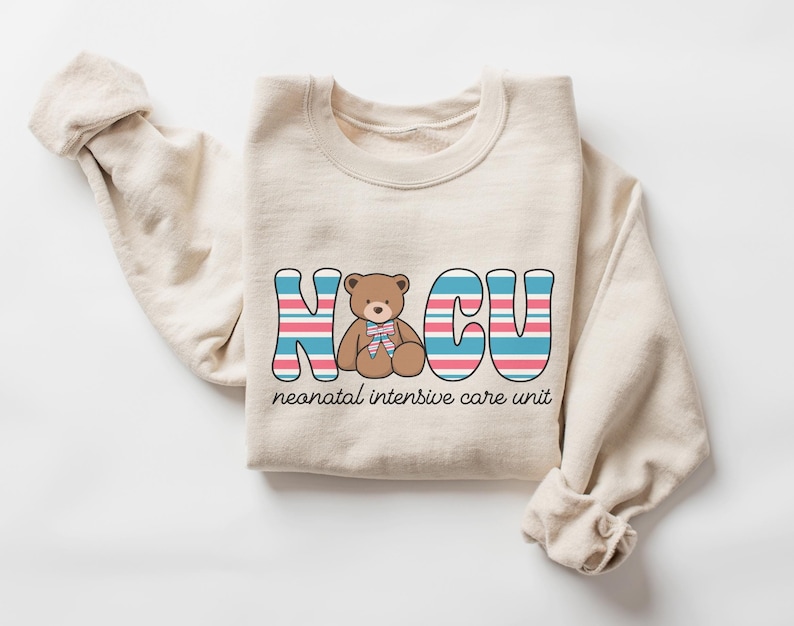 Puede incluir: Una sudadera beige con un dise&ntilde;o gr&aacute;fico de las letras "NICU" en rayas azules, rosas y blancas. Un oso de peluche marr&oacute;n con un lazo rosa est&aacute; sentado en el medio de las letras. El texto "unidad de cuidados intensivos neonatales" est&aacute; impreso debajo de las letras.