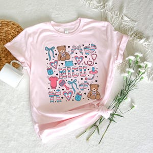 May include: Pink t-shirt with a graphic design of a teddy bear, a baby bottle, a rattle, a stethoscope, and the word "NICU".