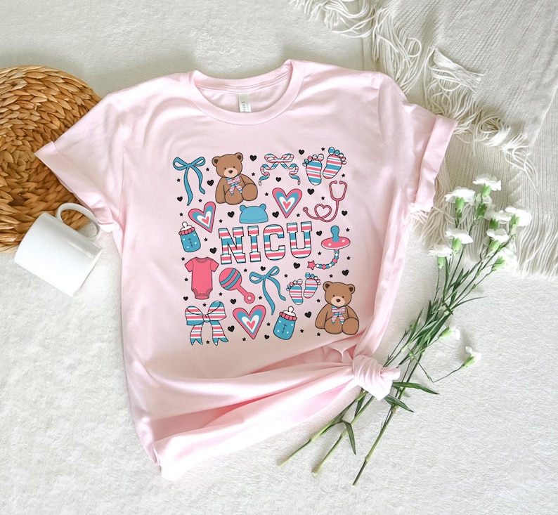 May include: Pink t-shirt with a graphic design of a teddy bear, a baby bottle, a rattle, a stethoscope, and the word "NICU".