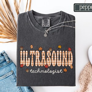 Puede incluir: Camiseta gris oscuro Comfort Colors con las palabras "ULTRASOUND" en un dise&ntilde;o a cuadros y "technologist" debajo. El dise&ntilde;o incluye hojas de oto&ntilde;o y bellotas. Tambi&eacute;n se ven un collar plateado y zapatos blancos.