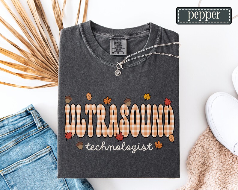 Puede incluir: Camiseta gris oscuro Comfort Colors con las palabras "ULTRASOUND" en un dise&ntilde;o a cuadros y "technologist" debajo. El dise&ntilde;o incluye hojas de oto&ntilde;o y bellotas. Tambi&eacute;n se ven un collar plateado y zapatos blancos.