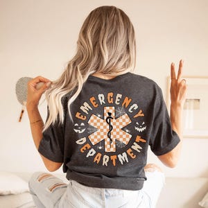 Op de afbeelding: Een donkergrijs t-shirt met een grote afbeelding op de achterkant met de tekst EMERGENCY DEPARTMENT in het oranje. Het ontwerp bevat een centraal oranje en wit geblokt symbool van de Ster van het Leven, spinnenwebben en twee pompoengezichten.