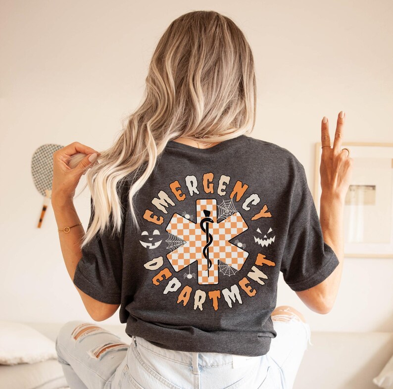 Op de afbeelding: Een donkergrijs t-shirt met een grote afbeelding op de achterkant met de tekst EMERGENCY DEPARTMENT in het oranje. Het ontwerp bevat een centraal oranje en wit geblokt symbool van de Ster van het Leven, spinnenwebben en twee pompoengezichten.