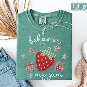 K&ouml;nnte beinhalten: Ein hellgr&uuml;nes T-Shirt mit Rundhalsausschnitt und einer Grafik aus drei Erdbeeren mit rot-wei&szlig;em Karomuster und gr&uuml;nen Vichy-Bl&auml;ttern. Der Text "behavior is my jam" ist in wei&szlig;er Schreibschrift gedruckt und von kleinen karierten Sternen umgeben.