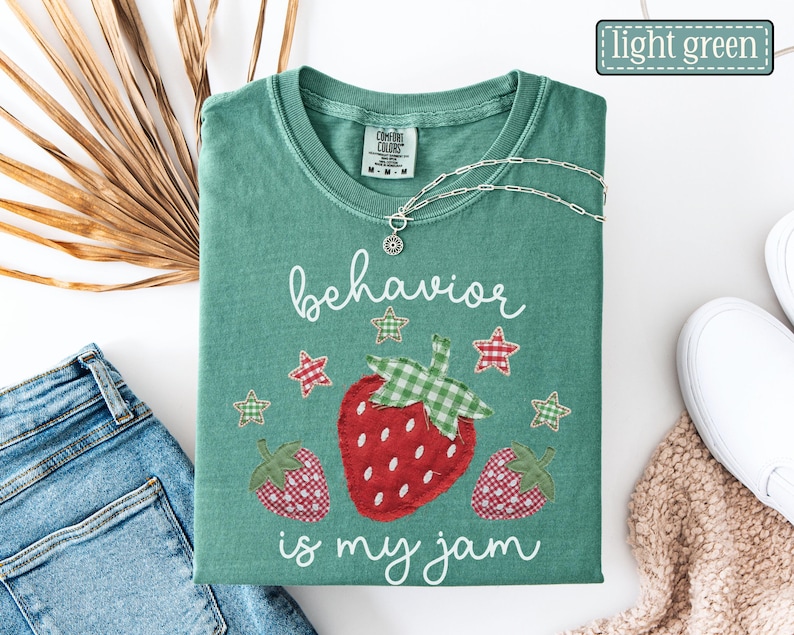 K&ouml;nnte beinhalten: Ein hellgr&uuml;nes T-Shirt mit Rundhalsausschnitt und einer Grafik aus drei Erdbeeren mit rot-wei&szlig;em Karomuster und gr&uuml;nen Vichy-Bl&auml;ttern. Der Text "behavior is my jam" ist in wei&szlig;er Schreibschrift gedruckt und von kleinen karierten Sternen umgeben.