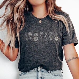 May include: A dark gray t-shirt with a white outline of a flower, a needle, and thread. The text "Roots" is written in white below the design.