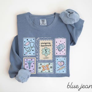 Pode incluir: Camisola azul jeans com gola redonda e mangas compridas. A frente apresenta seis patches retangulares com ilustra&ccedil;&otilde;es de tem&aacute;tica m&eacute;dica, incluindo um estetosc&oacute;pio, uma m&aacute;scara e uma ligadura. O texto "emergency department" tamb&eacute;m &eacute; vis&iacute;vel.