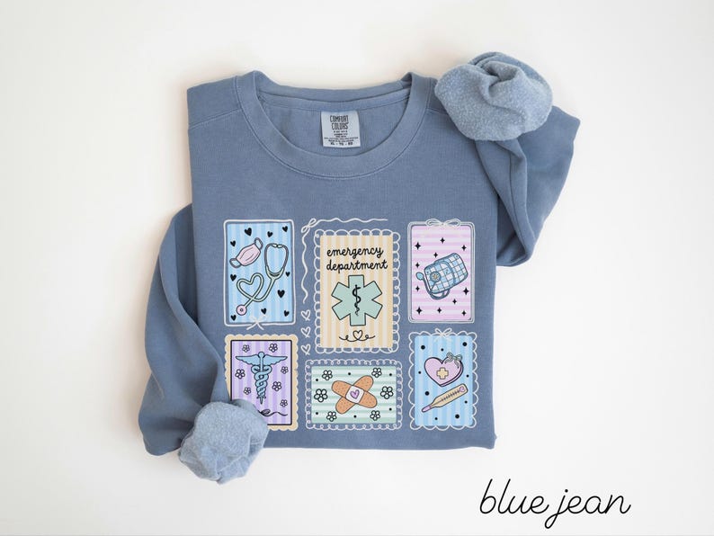 Pode incluir: Camisola azul jeans com gola redonda e mangas compridas. A frente apresenta seis patches retangulares com ilustra&ccedil;&otilde;es de tem&aacute;tica m&eacute;dica, incluindo um estetosc&oacute;pio, uma m&aacute;scara e uma ligadura. O texto "emergency department" tamb&eacute;m &eacute; vis&iacute;vel.