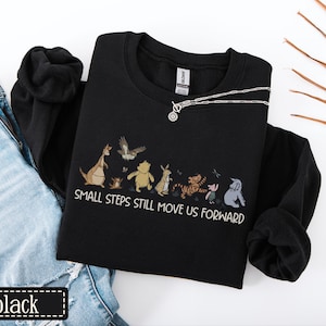 May include: Black sweatshirt with a graphic of cartoon characters and the text "SMALL STEPS STILL MOVE US FORWARD." A silver necklace with a circular pendant is on the sweatshirt. The word "black" is in a black rectangle.