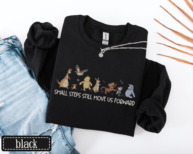 May include: Black sweatshirt with a graphic of cartoon characters and the text "SMALL STEPS STILL MOVE US FORWARD." A silver necklace with a circular pendant is on the sweatshirt. The word "black" is in a black rectangle.