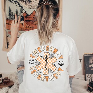 Op de afbeelding: Een wit t-shirt met een opdruk op de achterkant met de tekst EMERGENCY DEPARTMENT in het oranje. Het ontwerp bevat een centraal oranje en wit geblokt symbool van de Ster van het Leven met een zwarte esculaapstaf, spinnenwebben en spinnen.