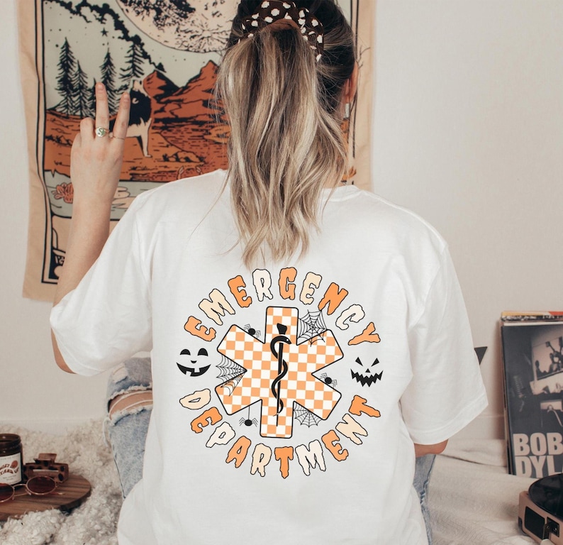 Op de afbeelding: Een wit t-shirt met een opdruk op de achterkant met de tekst EMERGENCY DEPARTMENT in het oranje. Het ontwerp bevat een centraal oranje en wit geblokt symbool van de Ster van het Leven met een zwarte esculaapstaf, spinnenwebben en spinnen.
