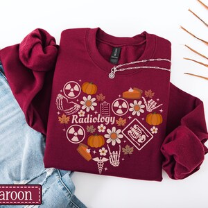 May include: A maroon crewneck sweatshirt featuring a heart-shaped graphic composed of pumpkins, autumn leaves, daisies, radiation symbols, a clipboard, a slice of pie, a medical caduceus, and skeletal hands. The word Radiology is printed in the center.