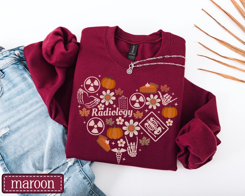 May include: A maroon crewneck sweatshirt featuring a heart-shaped graphic composed of pumpkins, autumn leaves, daisies, radiation symbols, a clipboard, a slice of pie, a medical caduceus, and skeletal hands. The word Radiology is printed in the center.