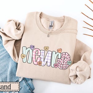 May include: A sand-colored sweatshirt with the word "neuro" in a colorful, playful font. The design includes a brain graphic and heart-shaped candies with phrases like "caffeine" and "brainy cupid". The sweatshirt is paired with blue jeans.