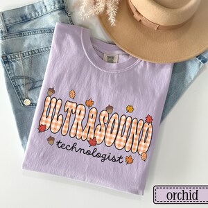 Puede incluir: Una camiseta de color lila con las palabras "ULTRASOUND technologist" en un dise&ntilde;o retro a cuadros naranja y blanco, adornada con hojas de oto&ntilde;o y bellotas. La camiseta est&aacute; doblada sobre unos vaqueros azules, con un sombrero beige y flores secas en el fondo.