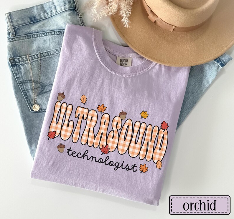 Puede incluir: Una camiseta de color lila con las palabras "ULTRASOUND technologist" en un dise&ntilde;o retro a cuadros naranja y blanco, adornada con hojas de oto&ntilde;o y bellotas. La camiseta est&aacute; doblada sobre unos vaqueros azules, con un sombrero beige y flores secas en el fondo.