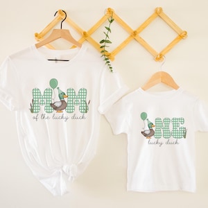 May include: White t-shirts with a green and white checkered design. The larger shirt says "Mom of the lucky duck" with a duck wearing a party hat and holding a balloon. The smaller shirt says "One lucky duck" with a duck wearing a party hat and holding a balloon.
