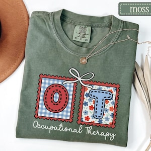 May include: Moss green t-shirt with the letters "OT" in red and blue postage stamp designs. The "O" has a red gingham pattern, and the "T" has a floral pattern. The words "Occupational Therapy" are printed below the letters.