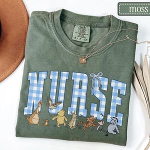 K&ouml;nnte beinhalten: Moosgr&uuml;nes T-Shirt mit dem Wort "NURSE" in blau-wei&szlig;em Vichydruck. Das Shirt zeigt Cartoon-Figuren am unteren Rand. Eine goldene Halskette mit Anh&auml;nger ist auf dem Shirt drapiert.