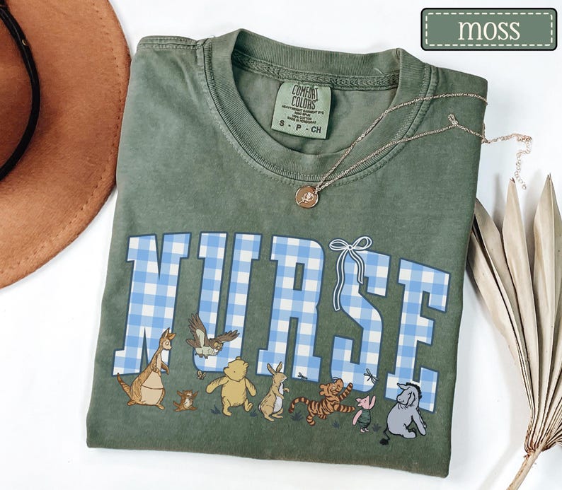K&ouml;nnte beinhalten: Moosgr&uuml;nes T-Shirt mit dem Wort "NURSE" in blau-wei&szlig;em Vichydruck. Das Shirt zeigt Cartoon-Figuren am unteren Rand. Eine goldene Halskette mit Anh&auml;nger ist auf dem Shirt drapiert.