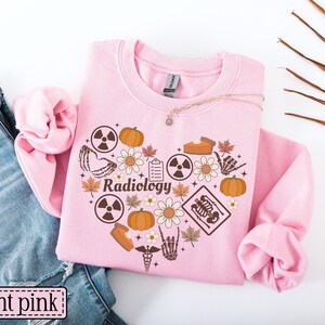 May include: A light pink crewneck sweatshirt featuring a heart-shaped graphic composed of brown icons including pumpkins, daisies, maple leaves, radiation symbols, a skeleton hand, a clipboard, a slice of pie, and the word Radiology in script.