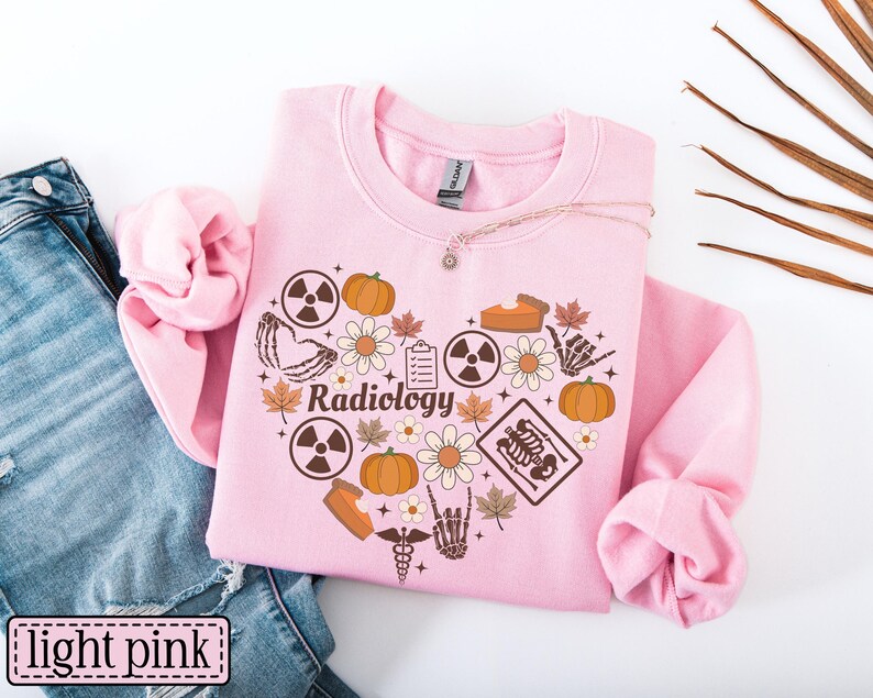 May include: A light pink crewneck sweatshirt featuring a heart-shaped graphic composed of brown icons including pumpkins, daisies, maple leaves, radiation symbols, a skeleton hand, a clipboard, a slice of pie, and the word Radiology in script.