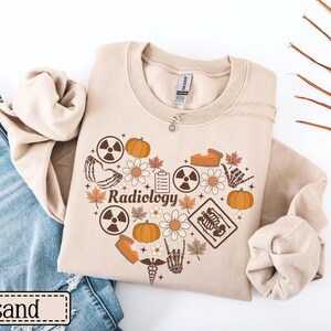 May include: A sand-colored crewneck sweatshirt featuring a heart-shaped graphic composed of pumpkins, autumn leaves, daisies, radiation symbols, a clipboard, a skeleton hand, and an X-ray image with the word Radiology printed in the center.