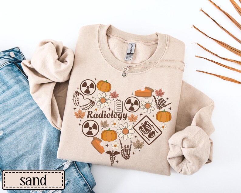 May include: A sand-colored crewneck sweatshirt featuring a heart-shaped graphic composed of pumpkins, autumn leaves, daisies, radiation symbols, a clipboard, a skeleton hand, and an X-ray image with the word Radiology printed in the center.