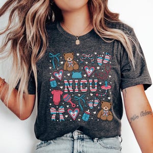 May include: A dark grey t-shirt with a colorful design featuring the word "NICU" surrounded by baby-themed illustrations, including teddy bears, footprints, bows, and a pacifier.