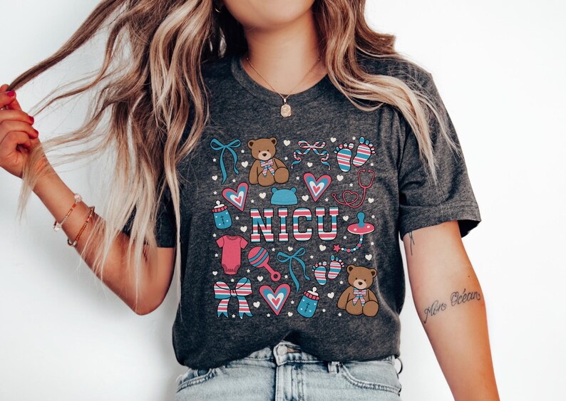 May include: A dark grey t-shirt with a colorful design featuring the word "NICU" surrounded by baby-themed illustrations, including teddy bears, footprints, bows, and a pacifier.