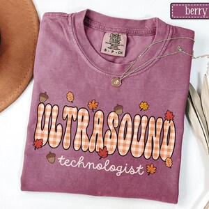 Puede incluir: Camiseta color bayas con la frase "ULTRASOUND technologist" en un estilo retro con un estampado de cuadros. El dise&ntilde;o incluye hojas de oto&ntilde;o y bellotas. La camiseta tiene cuello redondo y mangas cortas. Un collar dorado est&aacute; sobre la camiseta.